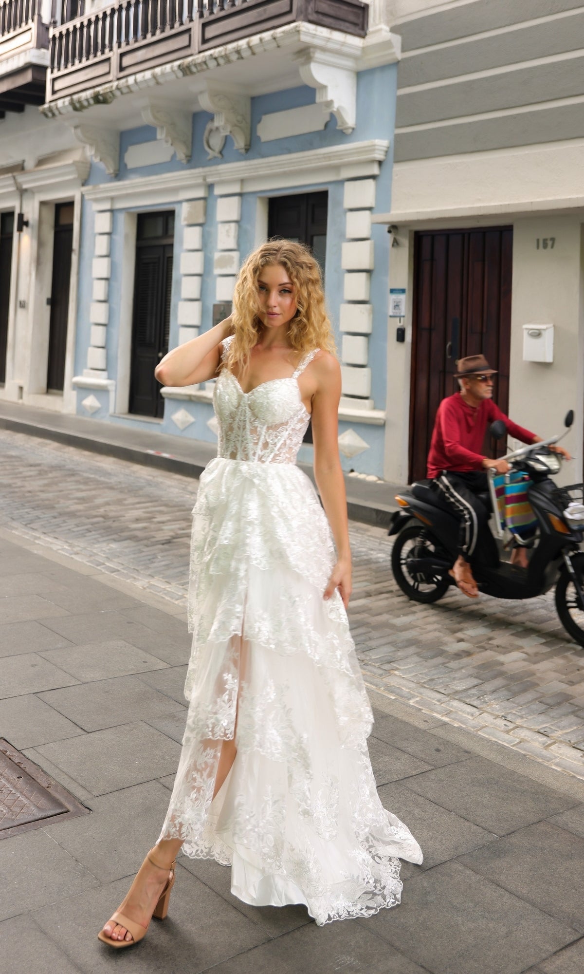 Wearing the Long Lace Ruffled A-Line Prom Dress T1335 and heels, a woman stands on a city sidewalk while a person on a scooter rides by in the background.